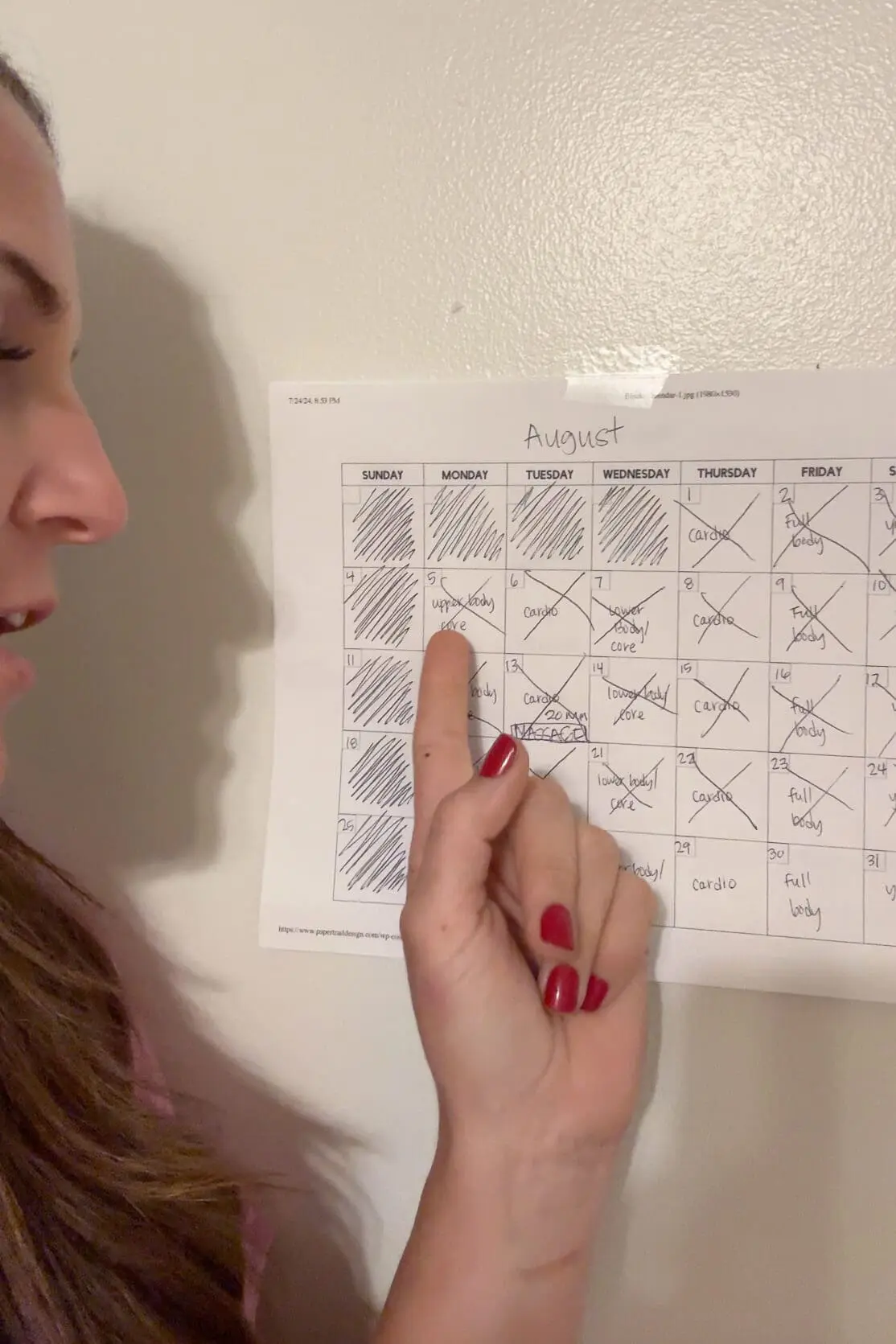 Close-up of a woman’s finger pointing to a day on her August workout calendar, which is filled with various exercises like cardio and strength training. This organized schedule highlights a structured workout routine for staying on track with fitness goals.